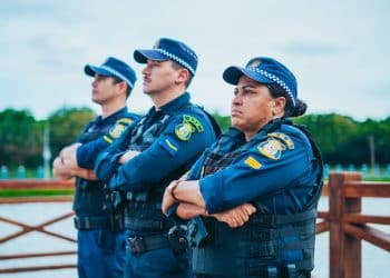 Três guardas municipais com coletes e uniformes azul-marinho olhando à frente com braços cruzados em área externa