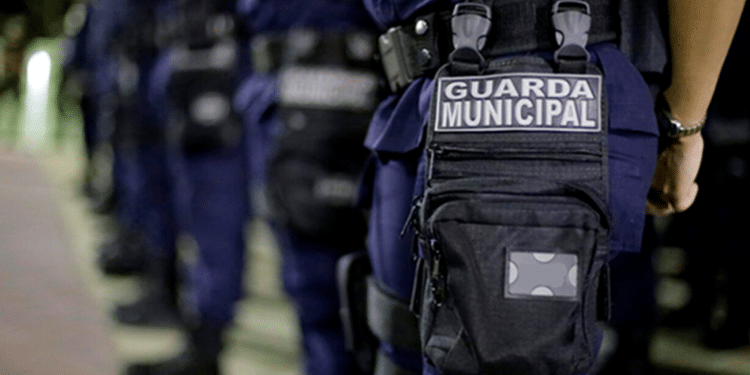 Policiais da Guarda Municipal alinhados usando uniformes azuis com bolsos e distintivos visíveis.