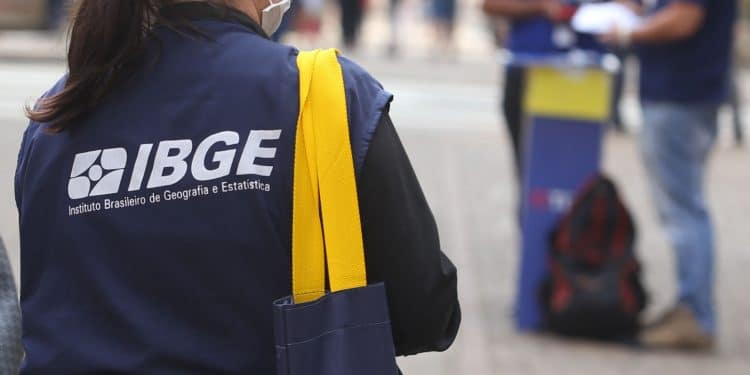 Mulher com colete do IBGE e máscara, carregando bolsa azul com alça amarela em área urbana