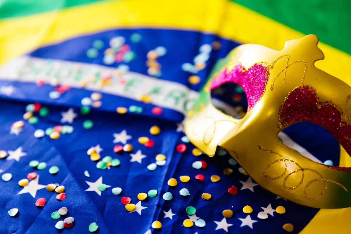 Máscara dourada e confetes representando o Carnaval no Brasil Máscara dourada com glitter sobre a bandeira do Brasil decorada com confetes, simbolizando as celebrações do Carnaval 2026.