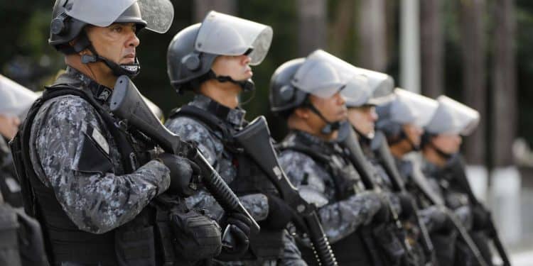 Policiais militares alinhados usando uniforme camuflado, capacete com visor e armamento tático.