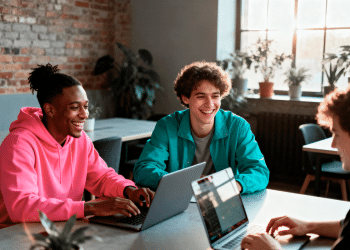 Três jovens homens sorrindo e trabalhando juntos em laptops em uma sala com paredes de tijolos e plantas ao fundo