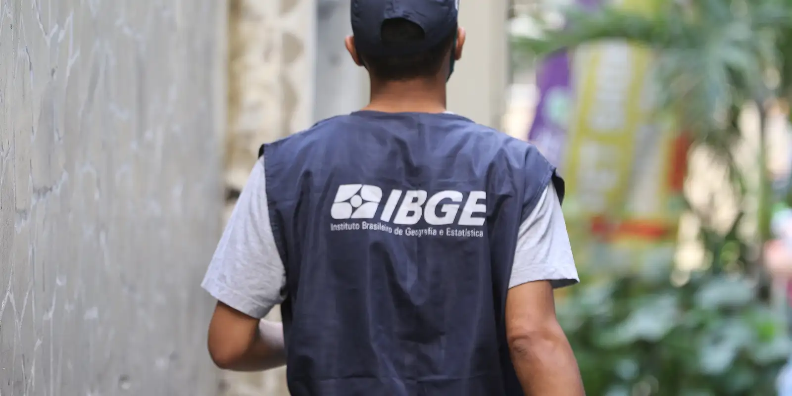 Vagas IBGE 2025 Agente do IBGE vestido com colete azul, de costas, realizando trabalho de campo em área urbana