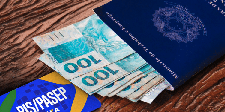 Carteira de trabalho com notas de cem reais e cartão PIS/PASEP da Caixa sobre mesa de madeira