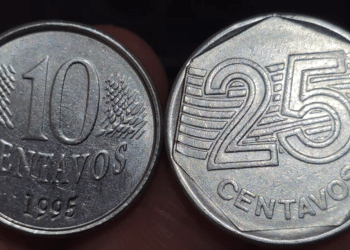 Grande quantidade de moedas de Real em destaque, com foco numa moeda de 25 centavos prateada ao centro e moedas de 1 Real douradas ao redor, evidenciando detalhes de relevo.