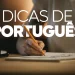Close das mãos de uma pessoa utilizando um lápis para escrever em um bloco de notas sobre uma mesa de madeira, com o título "Dicas de Português" em letras brancas grandes sobre a cena.
