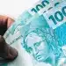 Nota de 100 reais do Brasil sendo segurada por uma mão, ilustrando o pagamento de aposentados do INSS em 2026.