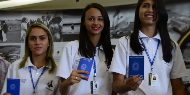 Três jovens sorridentes segurando carteira de trabalho azul e usando uniforme branco de trabalho
