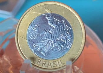 Macrofotografia de uma moeda de 1 Real das Olimpíadas Rio 2016, modalidade Judô. A imagem foca no detalhe do erro de cunhagem "data marcada" próximo ao joelho do atleta. A moeda tem borda dourada e centro prateado com a gravação de dois judocas em combate.