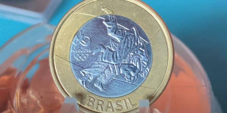 Macrofotografia de uma moeda de 1 Real das Olimpíadas Rio 2016, modalidade Judô. A imagem foca no detalhe do erro de cunhagem "data marcada" próximo ao joelho do atleta. A moeda tem borda dourada e centro prateado com a gravação de dois judocas em combate.