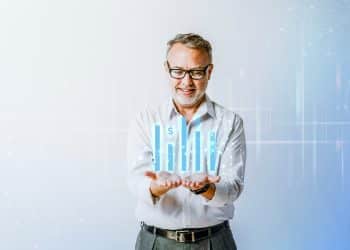 Homem sorridente com cabelos grisalhos e óculos segurando gráficos digitais de barras azuis com símbolos de dólar.