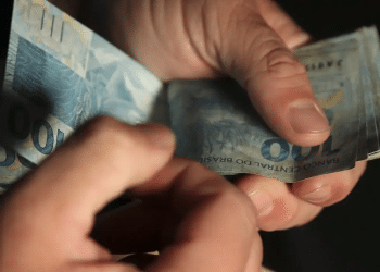 Mãos contando notas de R$ 100 reais referentes ao saque do bônus de R$ 1.000 do Pé-de-Meia em fevereiro