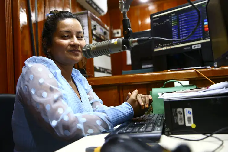 Mulher sorridente falando ao microfone em estúdio de rádio com painel de áudio e computador ao fundo
