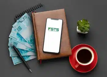 Celular com aplicativo FGTS sobre agenda de couro ao lado de notas de cem reais caneta e xícara de café.