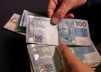 Close das mãos de uma pessoa segurando e contando diversas notas de cem reais (100 BRL) sobre uma superfície escura.