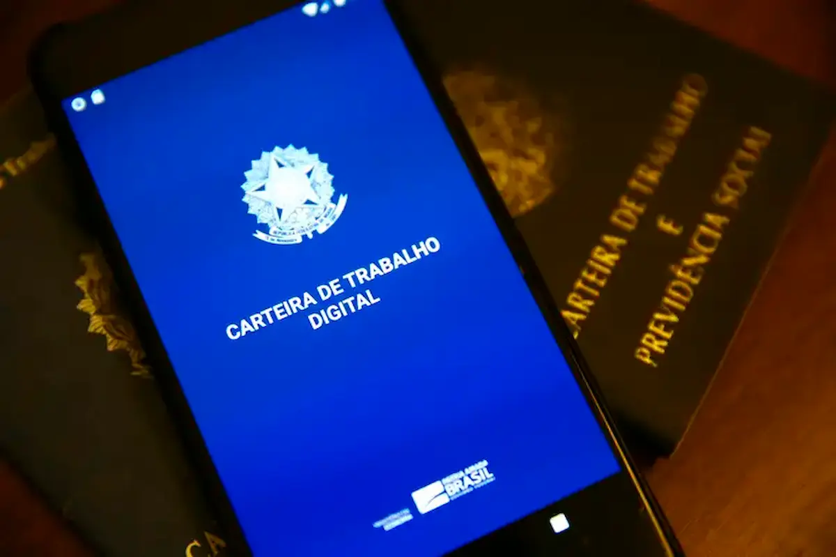 Imagem de um smartphone mostrando a Carteira de Trabalho Digital, com a versão física da carteira ao fundo, representando a discussão sobre a escala 6x1 no Brasil.