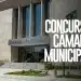 Fachada moderna da Câmara Municipal com bandeira do Brasil e anúncio de concurso público 2026