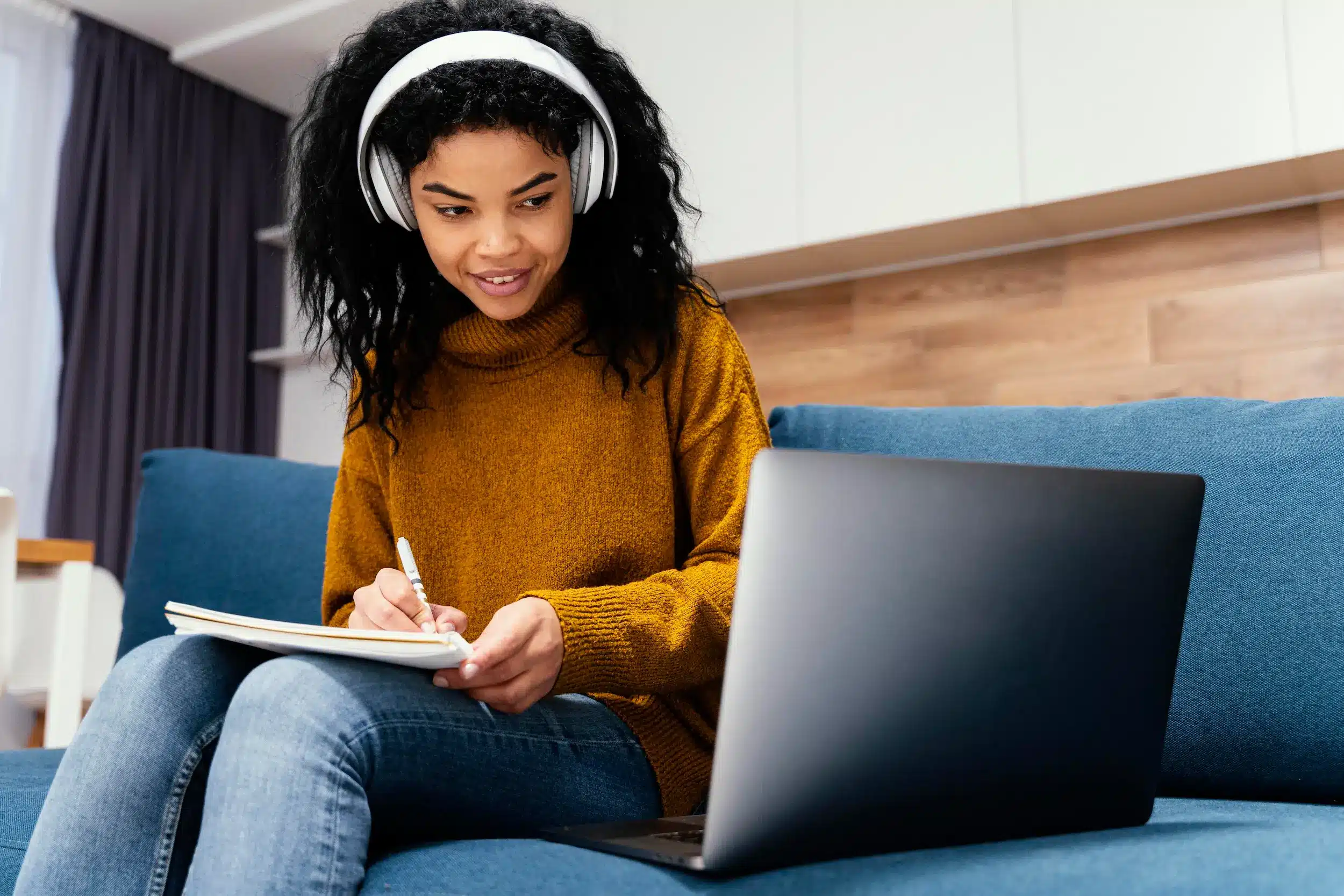Mulher com fones de ouvido, estudando e anotando em caderno enquanto usa o computador, ilustrando a flexibilidade dos cursos online.