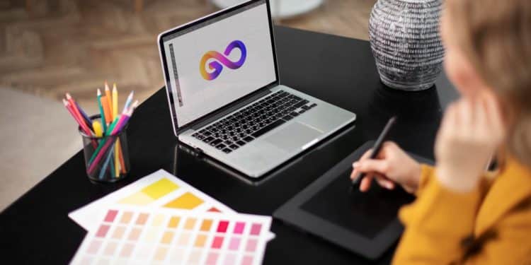 Vista superior de uma designer trabalhando em um laptop que exibe um logotipo colorido em formato de infinito, com uma mesa digitalizadora, amostras de cores e lápis ao redor.