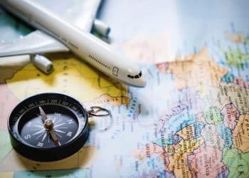 Visão aérea de um mapa-múndi colorido sobre o qual repousam uma bússola preta clássica e a miniatura de um avião comercial branco decolando sobre a Europa e o Mediterrâneo.