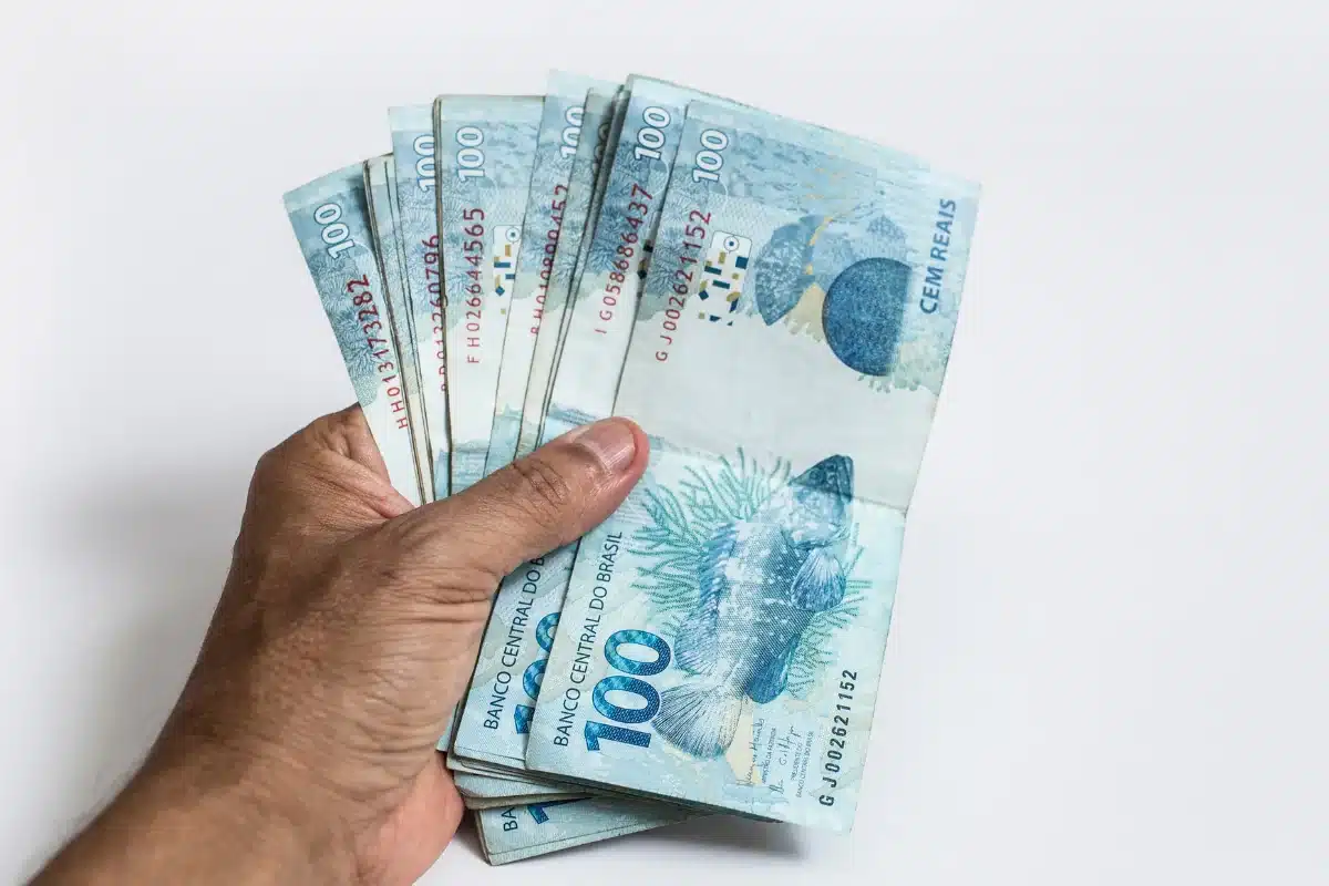 Mão masculina segurando um punhado de notas de cem reais em fundo branco