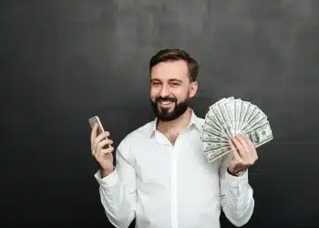 Homem sorrindo enquanto segura um maço de dinheiro e um celular, simbolizando a conquista de um bom salário com oportunidades que não exigem diploma superior.