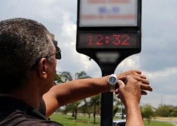 Homem de costas ajustando o relógio de pulso em frente a um termômetro de rua digital que marca 12:32 sob um céu ensolarado.