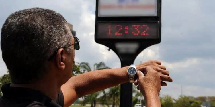 Homem de costas ajustando o relógio de pulso em frente a um termômetro de rua digital que marca 12:32 sob um céu ensolarado.