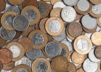 Fotografia em plano fechado de um monte de moedas de Real espalhadas, incluindo moedas de 1 Real, 50 centavos, 10 centavos e 5 centavos. As peças apresentam diferentes anos de cunhagem e estados de conservação, com destaque para o brilho do metal e detalhes do relevo.