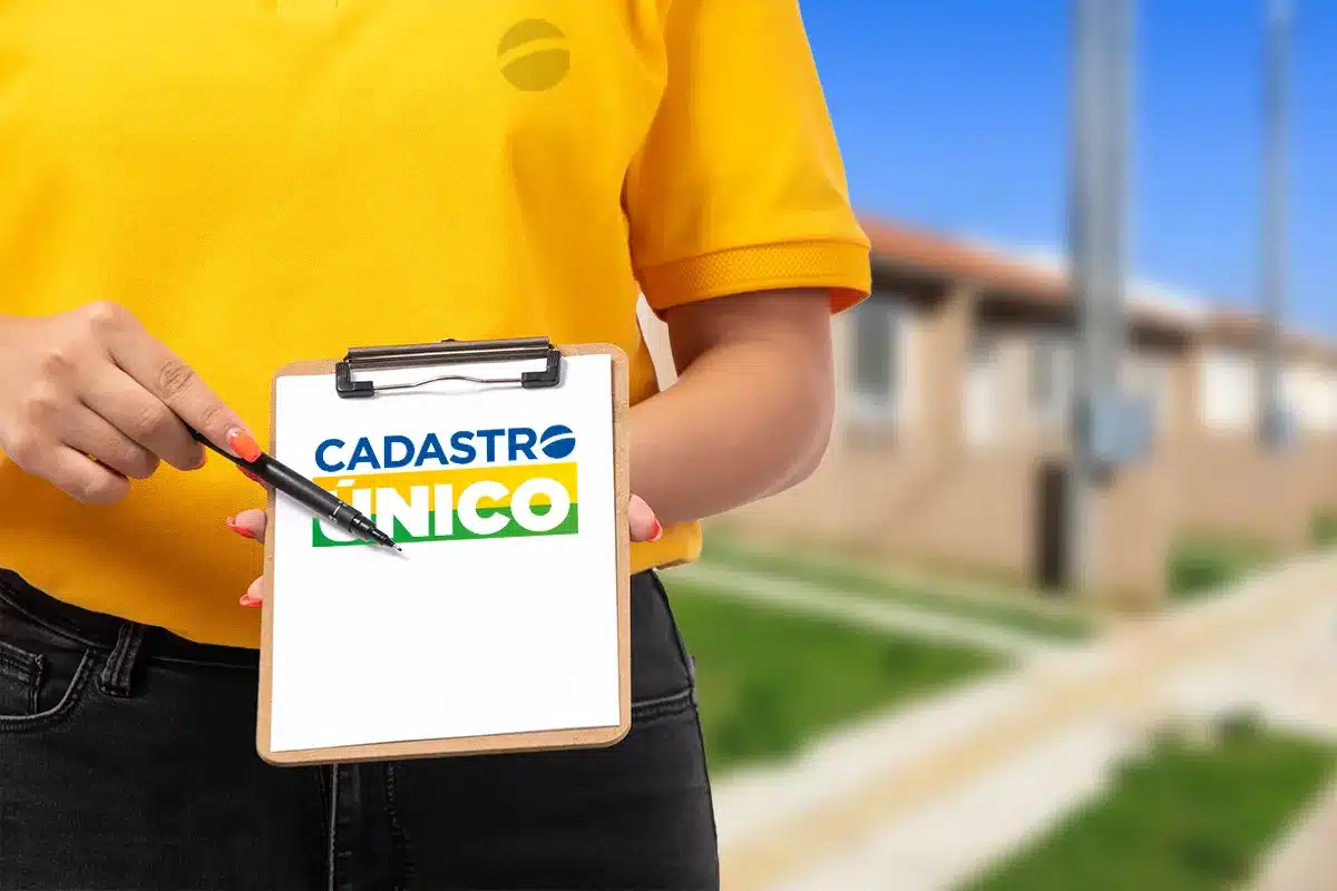 Close de uma pessoa vestindo camisa amarela, segurando uma prancheta de madeira. Na folha branca, destaca-se o logotipo oficial do Cadastro Único nas cores azul, amarelo e verde. Ao fundo, uma área residencial com casas populares está levemente desfocada.