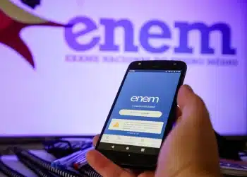 Mão segura celular aberto no app ENEM; fundo exibe logo ENEM em tela brilhante