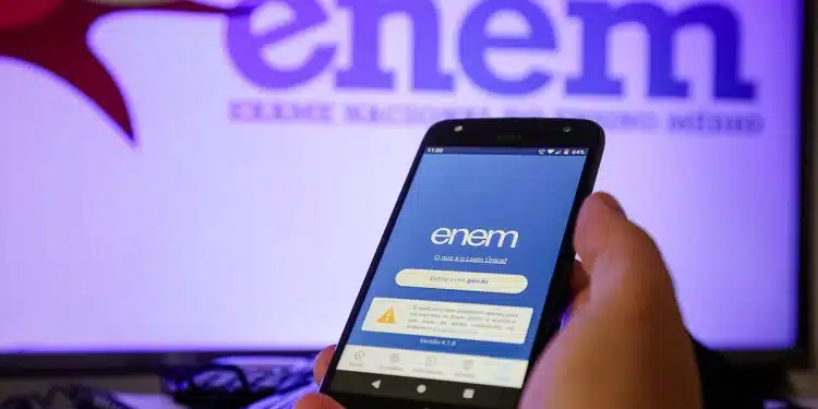 Mão segura celular aberto no app ENEM; fundo exibe logo ENEM em tela brilhante