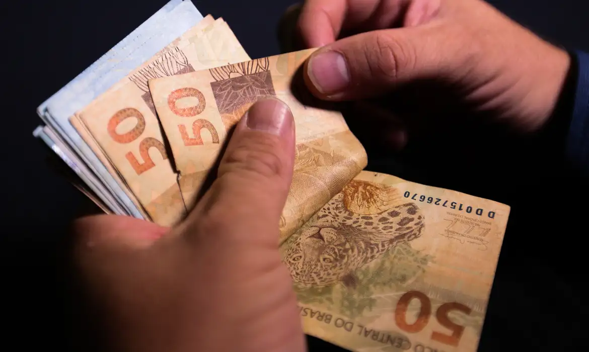 Close de mãos humanas segurando e folheando um maço de notas de 50 reais (BRL), com foco na textura do papel-moeda e na estampa da onça-pintada.