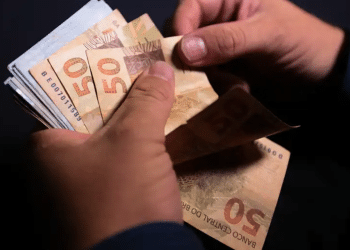 Mãos segurando várias notas de dinheiro em reais, especialmente notas de cinquenta reais.