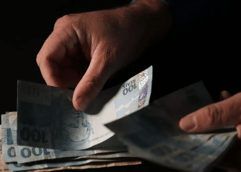 Mãos separando cédulas de 100 e 50 reais em fundo escuro representando pagamento do INSS acima do salário mínimo