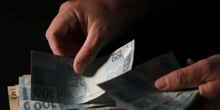 Mãos separando cédulas de 100 e 50 reais em fundo escuro representando pagamento do INSS acima do salário mínimo