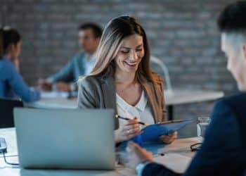 Executiva sorridente analisando documentos em reunião de trabalho com notebook e colegas ao fundo.