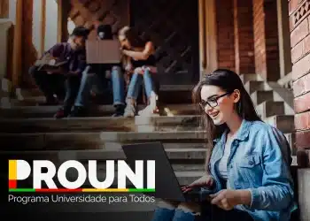 Estudante sorrindo enquanto trabalha no computador em um ambiente acadêmico, com outros estudantes ao fundo, em uma escada de um prédio.