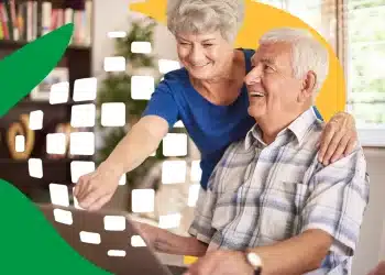 Casal de idosos sorrindo enquanto consulta computador em casa com logo do INSS ao fundo.