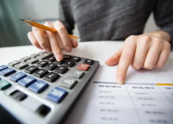 Pessoa utilizando uma calculadora para realizar cálculos financeiros, com uma planilha ao lado.