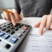 Pessoa utilizando uma calculadora para realizar cálculos financeiros, com uma planilha ao lado.