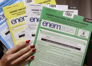 Mão segurando cadernos coloridos do ENEM 2025, amarelo, azul, branco, verde, para prova do 1º dia.