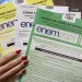 Mão segurando cadernos coloridos do ENEM 2025, amarelo, azul, branco, verde, para prova do 1º dia.