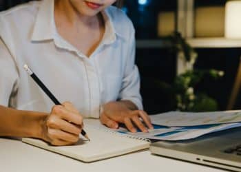 Close das mãos de uma revisora de textos escrevendo anotações em um caderno, com folhas de rascunho e notebook sobre a mesa em ambiente calmo.