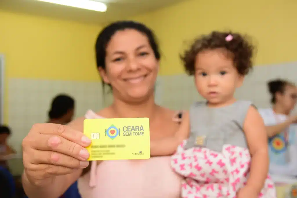 Mulher segura o Cartão Ceará Sem Fome enquanto carrega uma criança no colo.