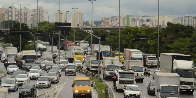 Vista aérea de uma rodovia movimentada com diversos carros, caminhões e vans em circulação sob um céu nublado, com prédios e vegetação ao fundo.
