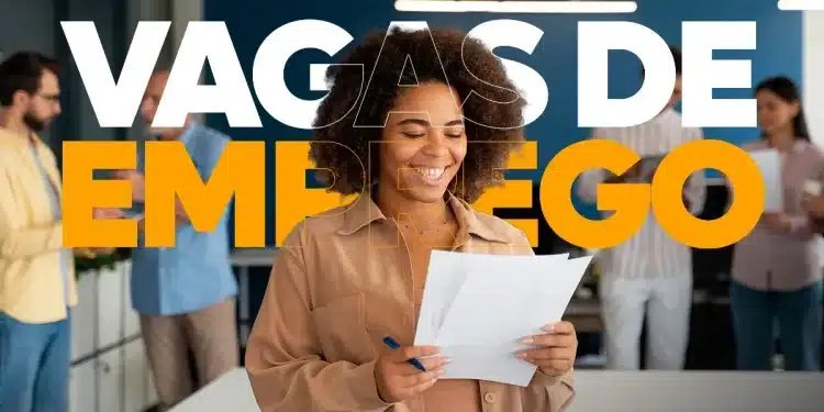 Mulher sorridente segurando documentos de emprego, com equipe ao fundo em ambiente corporativo.