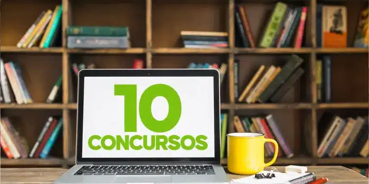 Laptop em mesa exibe “10 concursos” ao lado de caneca amarela e cadernos, com estante de livros ao fundo.