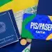 Carteira de trabalho, notas de cem reais, moedas e cartão PIS/Pasep sobre bandeira do Brasil.