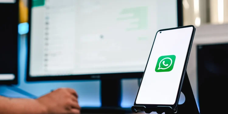 Celular exibindo o logo do WhatsApp em frente a dois monitores de computador. A tela de fundo exibe uma interface de chat do WhatsApp.